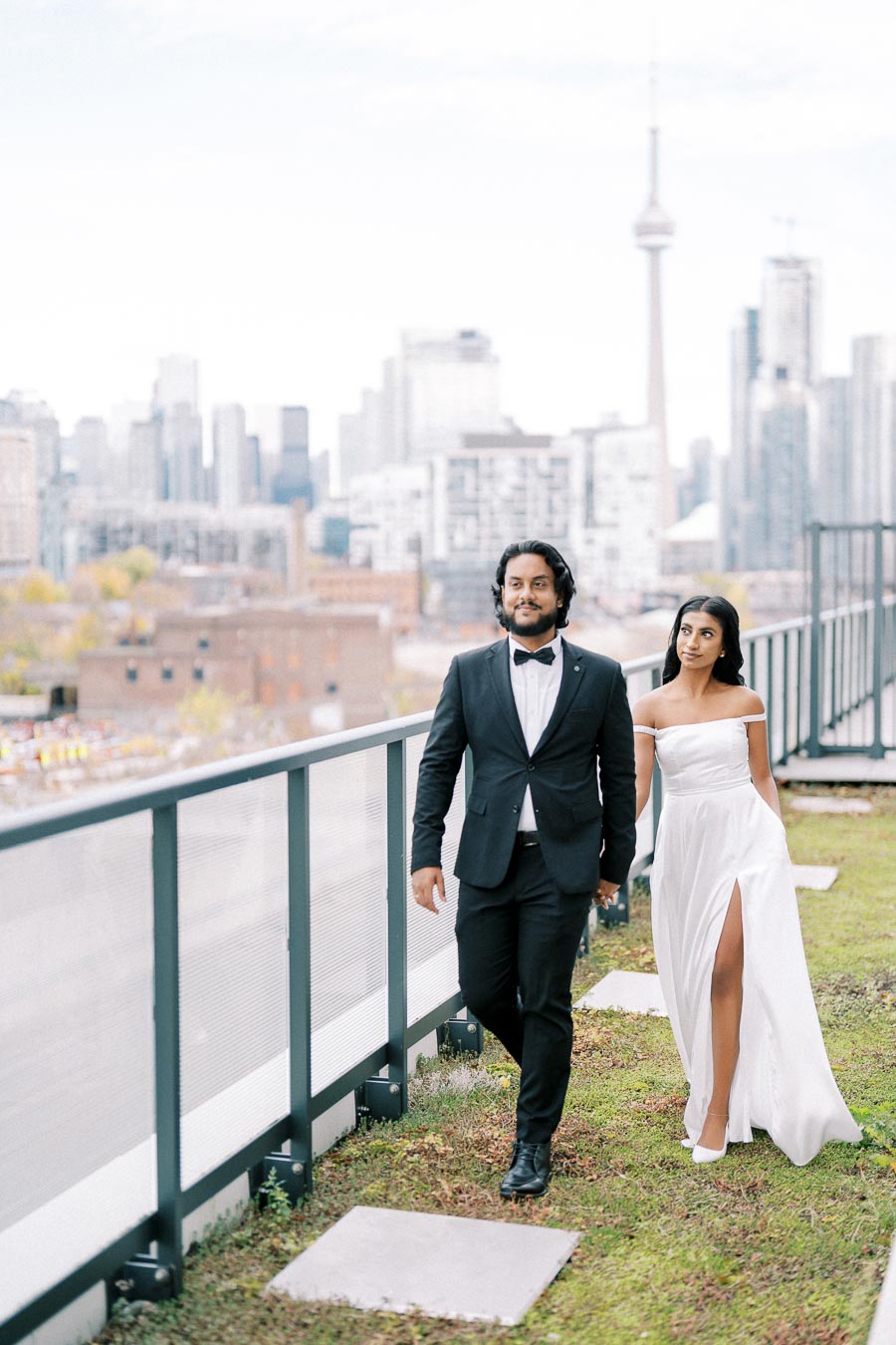 Couple in elegant attire walking on a rooftop garden with a city skyline in the background