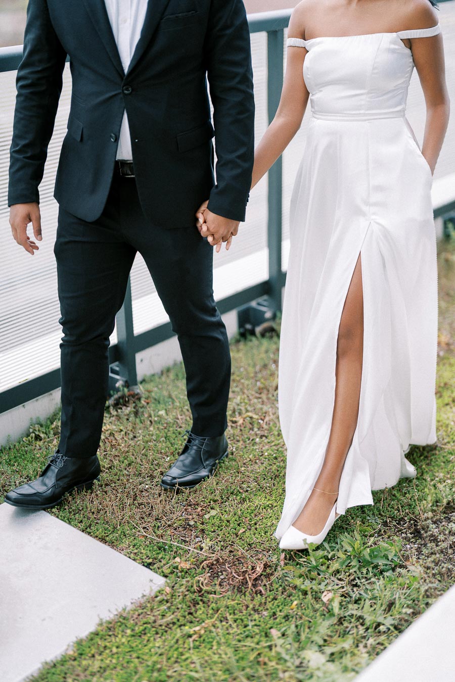 Couple holding hands in formal attire, with the woman in a white gown featuring a thigh-high slit, and the man in a black suit on a grassy path.