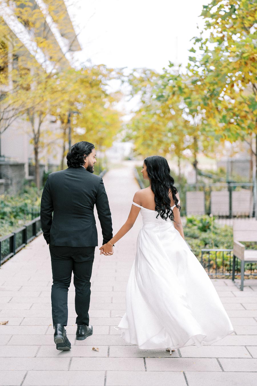 Bride and groom walking hand in hand on a tree-lined pathway, with the bride wearing a white gown and the groom in a black suit, surrounded by autumn foliage.