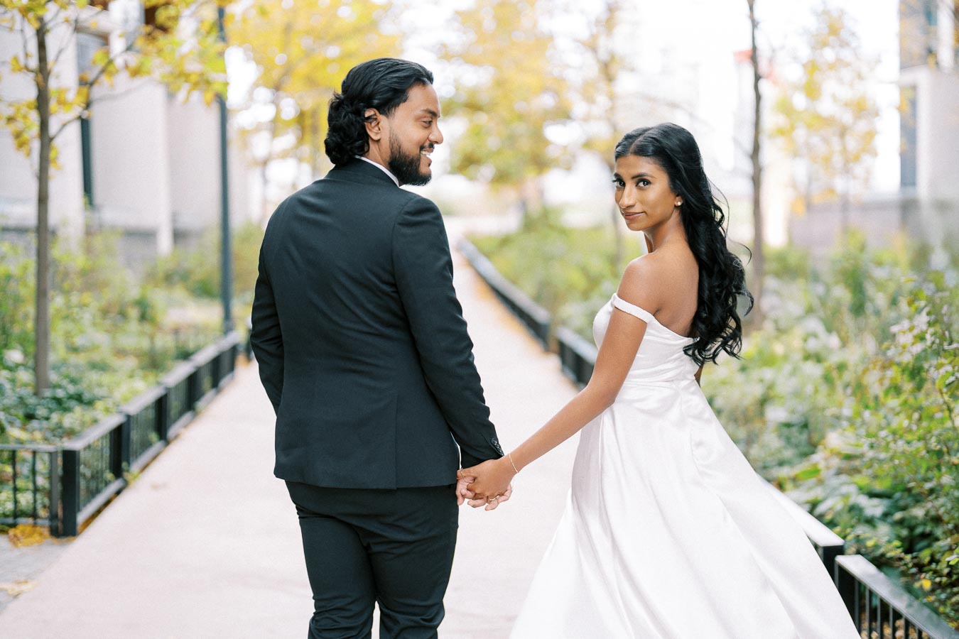 A happy couple walking hand in hand in a scenic outdoor setting, with the woman wearing an elegant white dress and the man in a sleek black suit, surrounded by lush greenery and soft golden light.