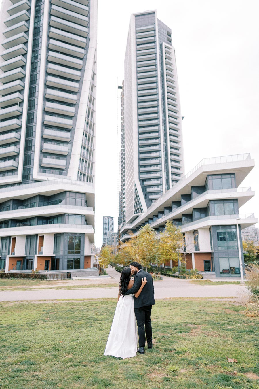 A couple embraces while walking towards modern high-rise apartment buildings, surrounded by a landscaped park setting.