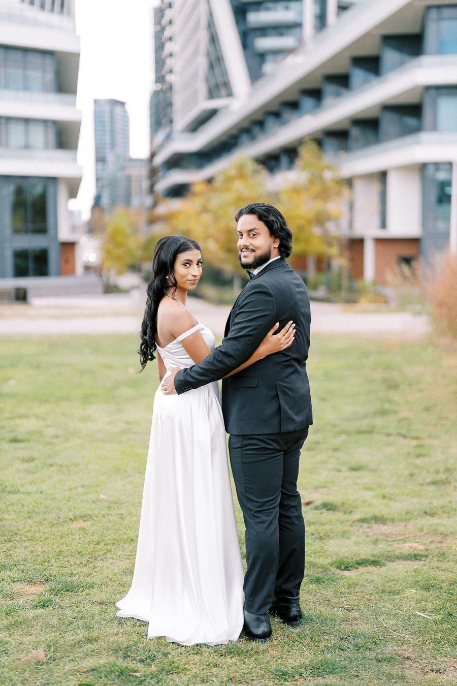 Couple embracing in front of modern architecture outdoors, woman in white dress and man in black suit, with green grass and trees in the background.