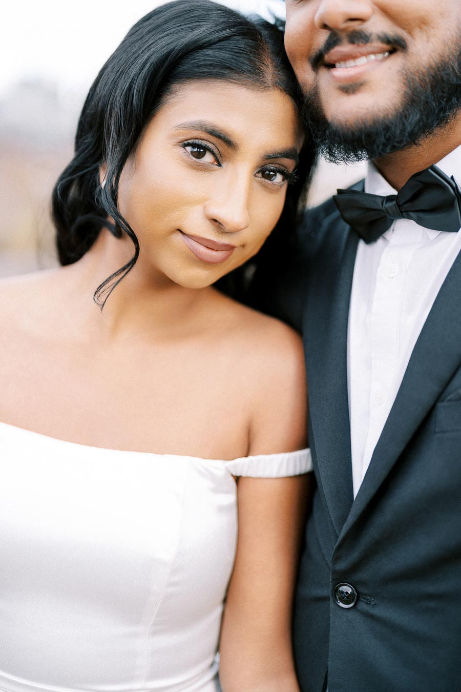 A couple dressed in formal attire, with the woman in an off-the-shoulder white dress and the man in a black suit with a bow tie, smiling and posing closely together.