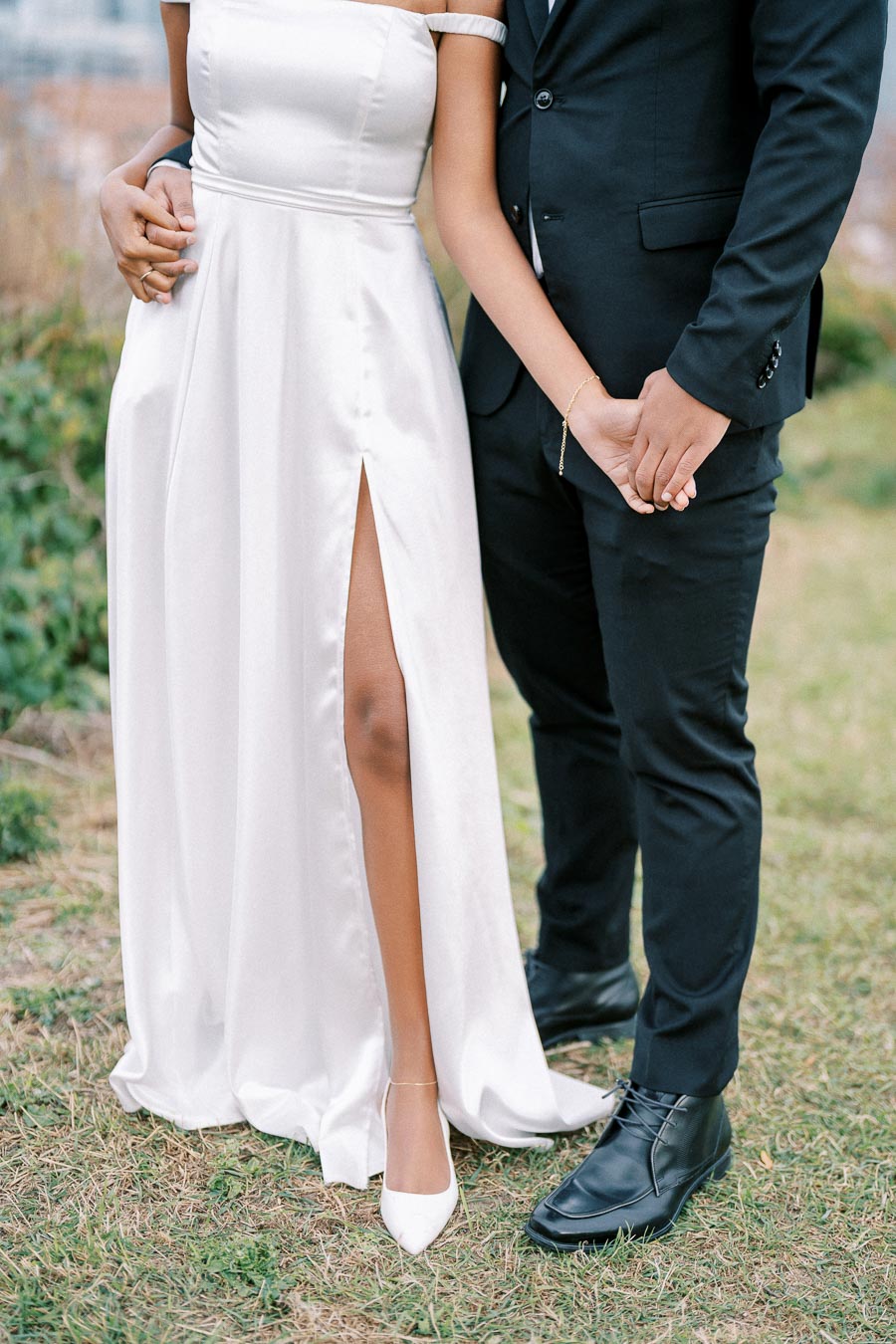 Couple Holding Hands in Elegant Attire, Bride in White Satin Dress with Slit and Groom in Black Suit, Outdoor Wedding Setting