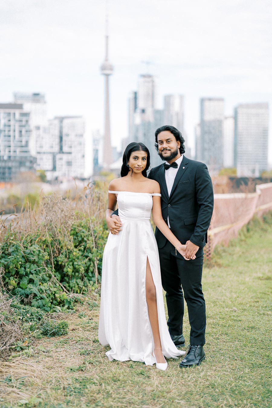 A couple dressed elegantly in formal attire posing outdoors with a city skyline in the background, featuring a prominent tall tower.