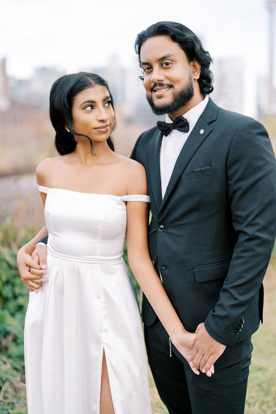 Elegant couple posing outdoors in formal attire, with the woman in a white strapless gown and the man in a black suit with a bow tie, holding hands and smiling.