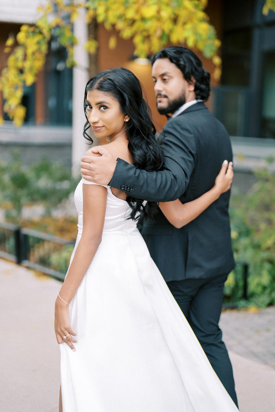 Elegant couple in formal attire embracing outdoors, with the woman wearing a white dress and the man in a dark suit, set against a backdrop of autumn foliage.