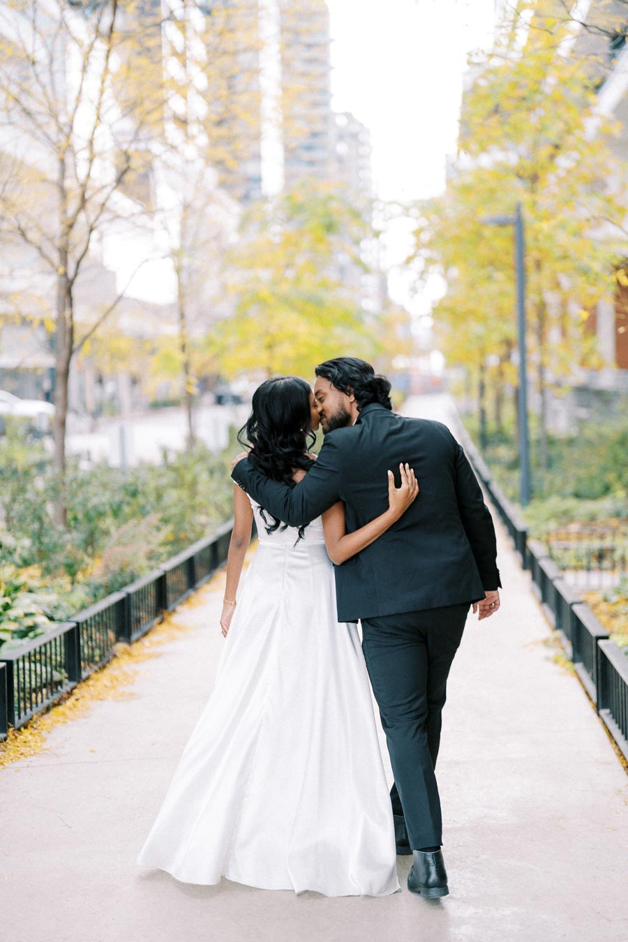 A newlywed couple walking hand in hand down a city path, the bride in a white dress, the groom in a black suit, surrounded by trees with autumn foliage.