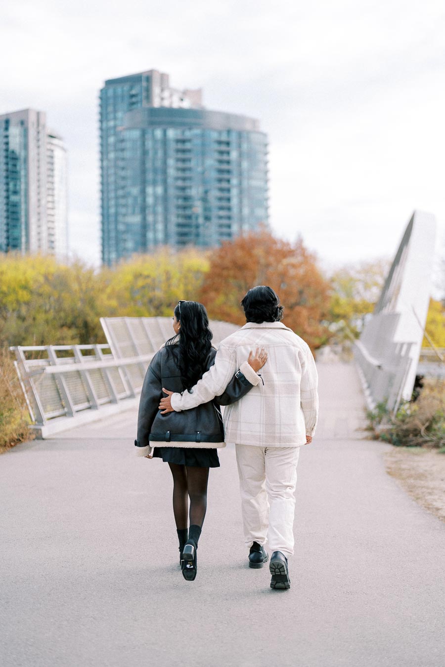 A couple walking arm-in-arm on a pathway with tall city buildings in the background, surrounded by autumn trees and a clear sky.
