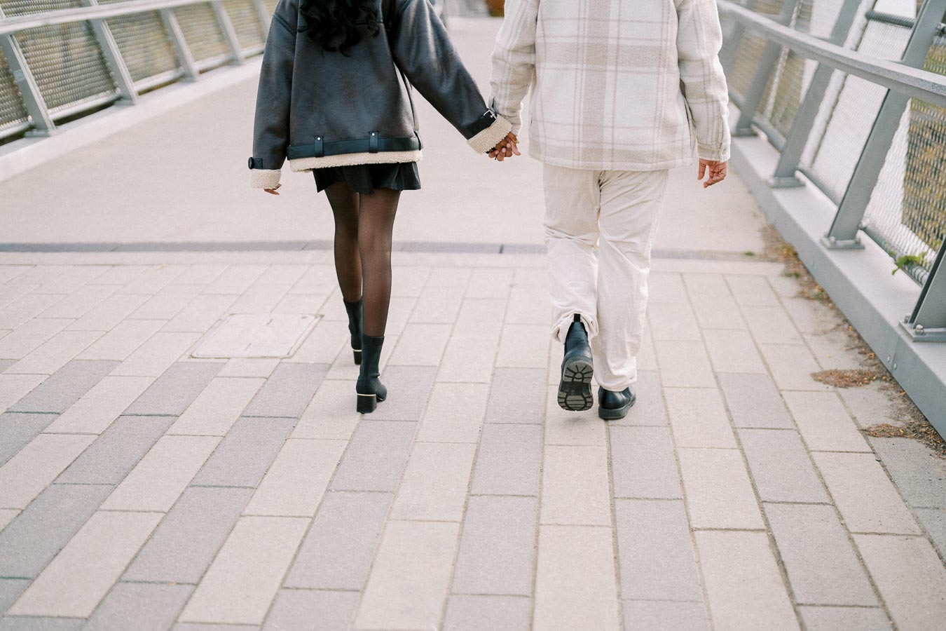 A couple walking hand in hand on a modern pedestrian bridge, wearing stylish winter clothing, with a focus on their outerwear and fashion-forward footwear.