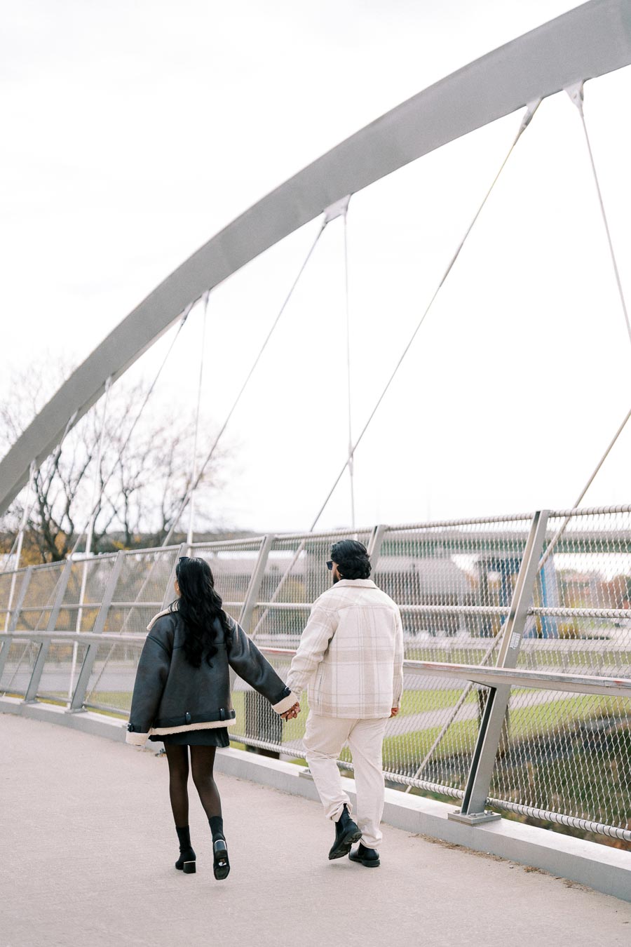 Couple holding hands while walking on a modern arched bridge, wearing stylish winter coats, showcasing urban romance and fashion.