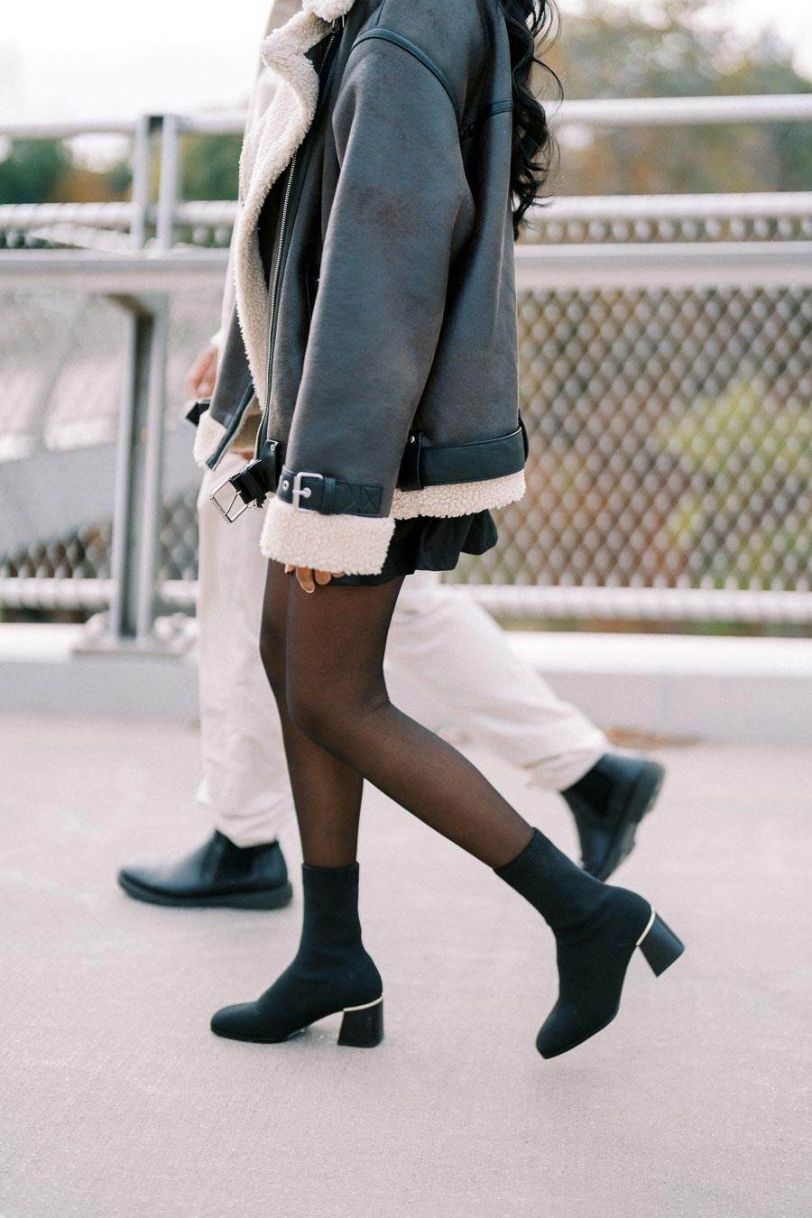 A person in a stylish black shearling jacket and black heeled boots walks on a concrete pathway, with a background of a metal fence and greenery. Fashionable autumn or winter outfit concept.