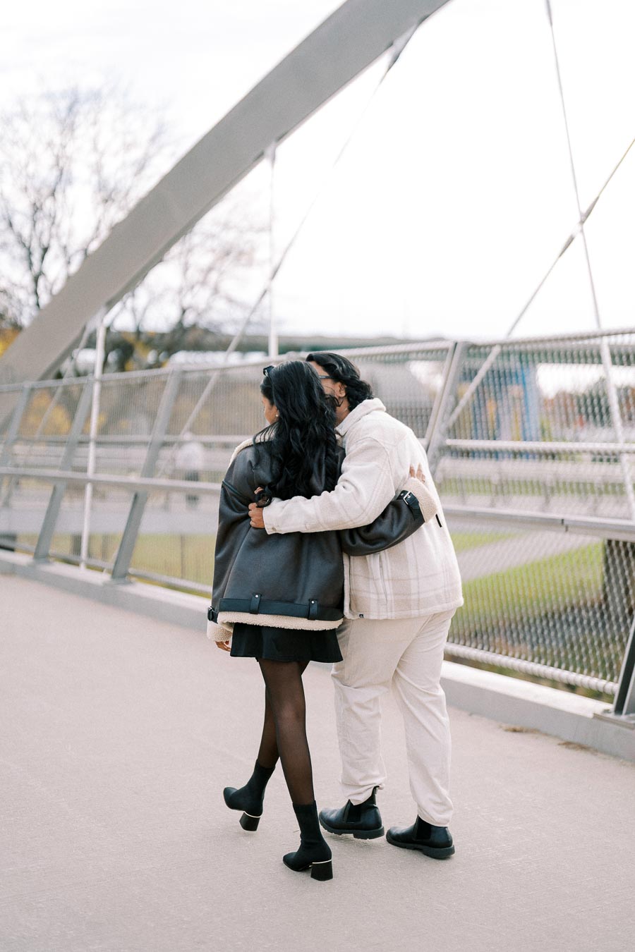 Couple walking on a bridge embracing each other, wearing stylish winter jackets, against an urban backdrop.