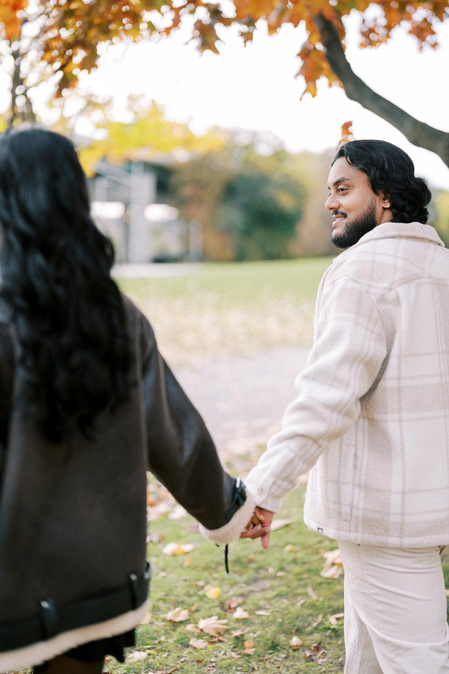 A couple holding hands and walking in a park during autumn with colorful leaves on the trees in the background.