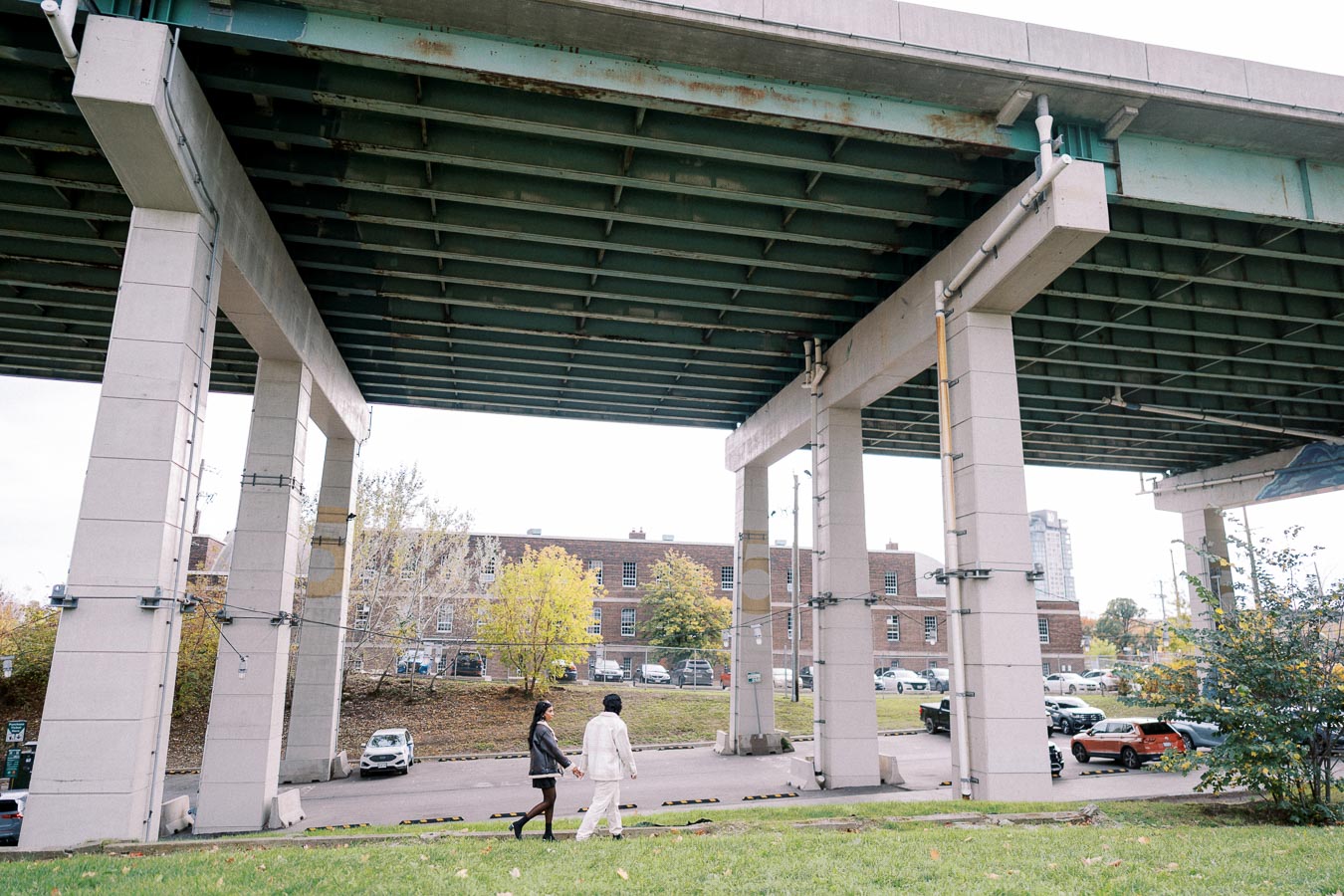 Two people walking hand in hand under a large overpass, with a backdrop of urban buildings and parked cars. The scene captures an urban setting with contrasting elements of greenery and infrastructure.