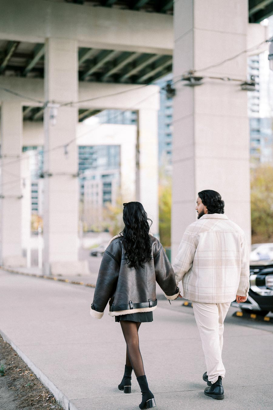 A couple walking hand in hand under an urban overpass, with modern city buildings in the background, both wearing stylish winter coats.