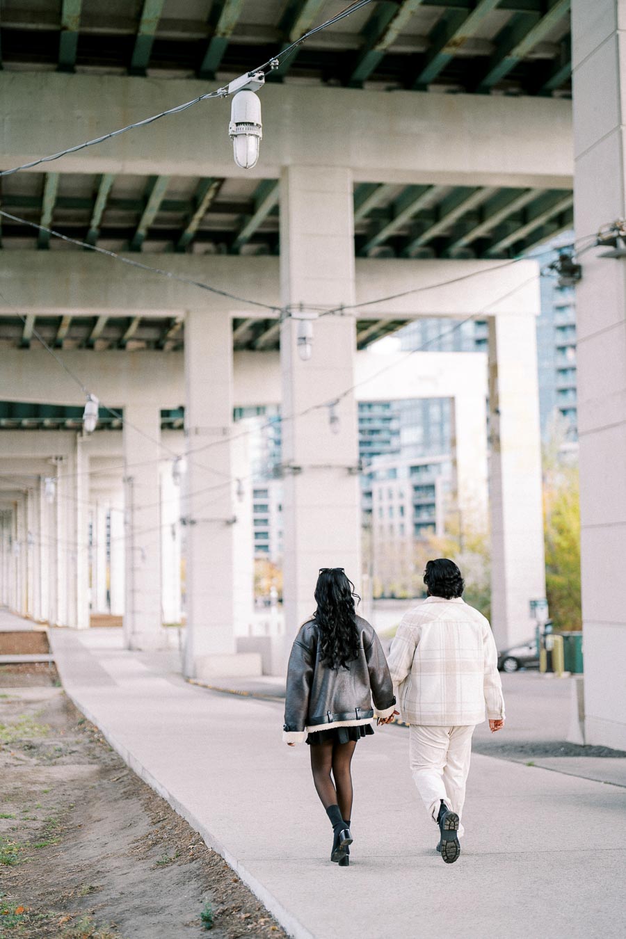 A couple walking hand in hand under an urban overpass, wearing stylish winter clothing, with city buildings in the background, daytime scene.