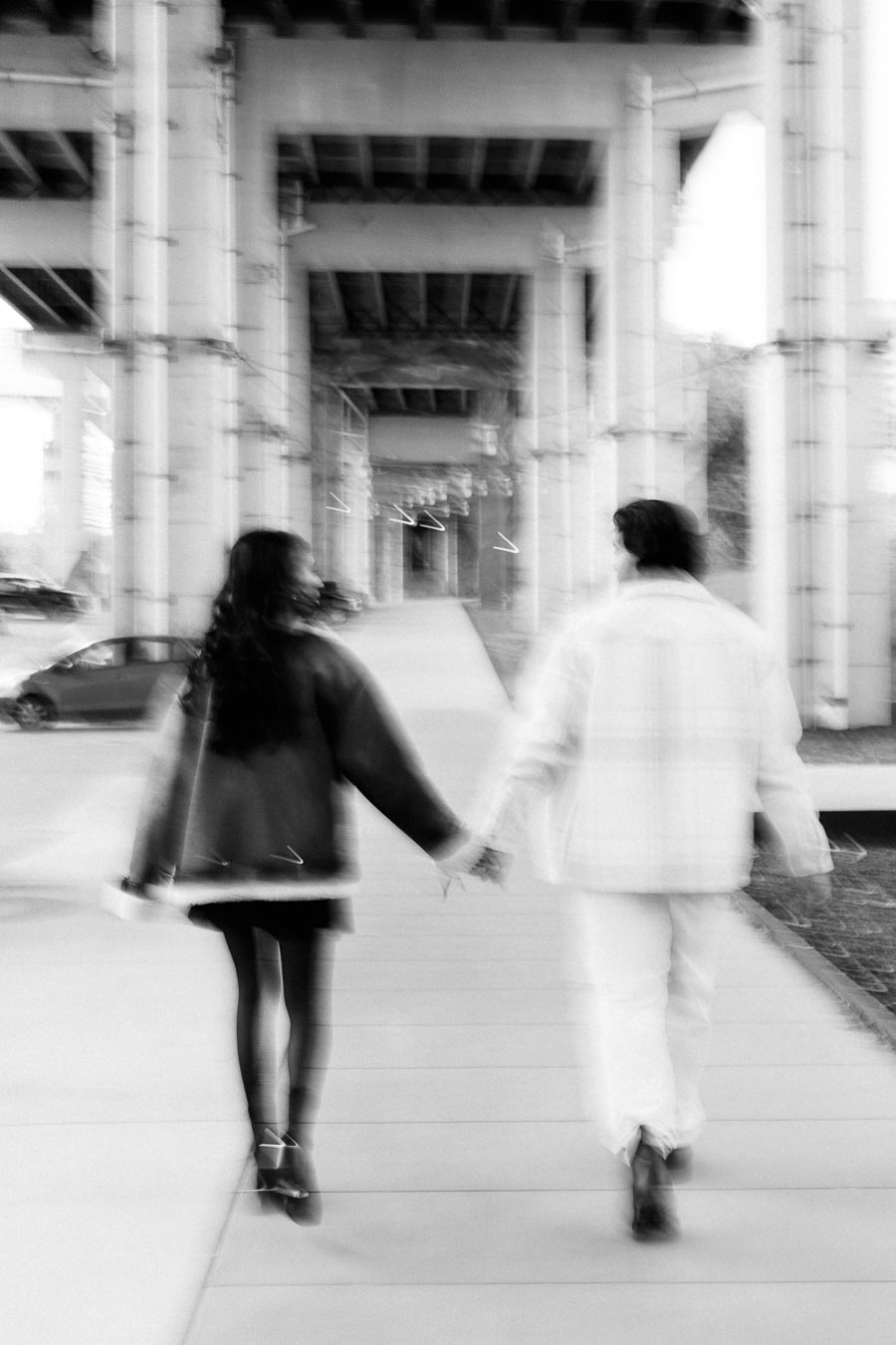 A black and white image of a couple walking hand in hand under a bridge, showcasing urban romance and architectural symmetry.