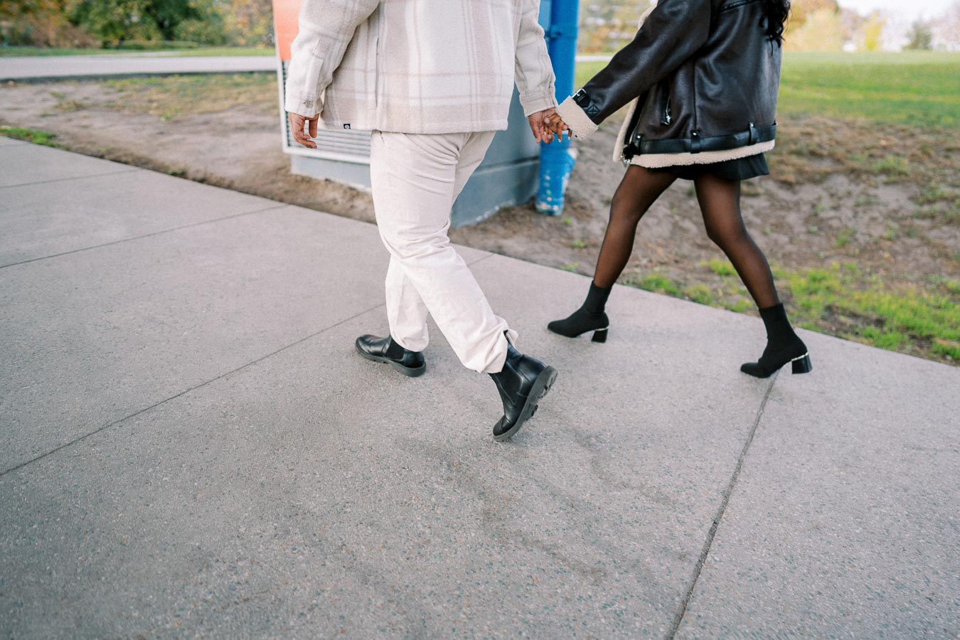 A couple holding hands while walking on a paved path, wearing stylish black and white outfits, with greenery visible in the background.