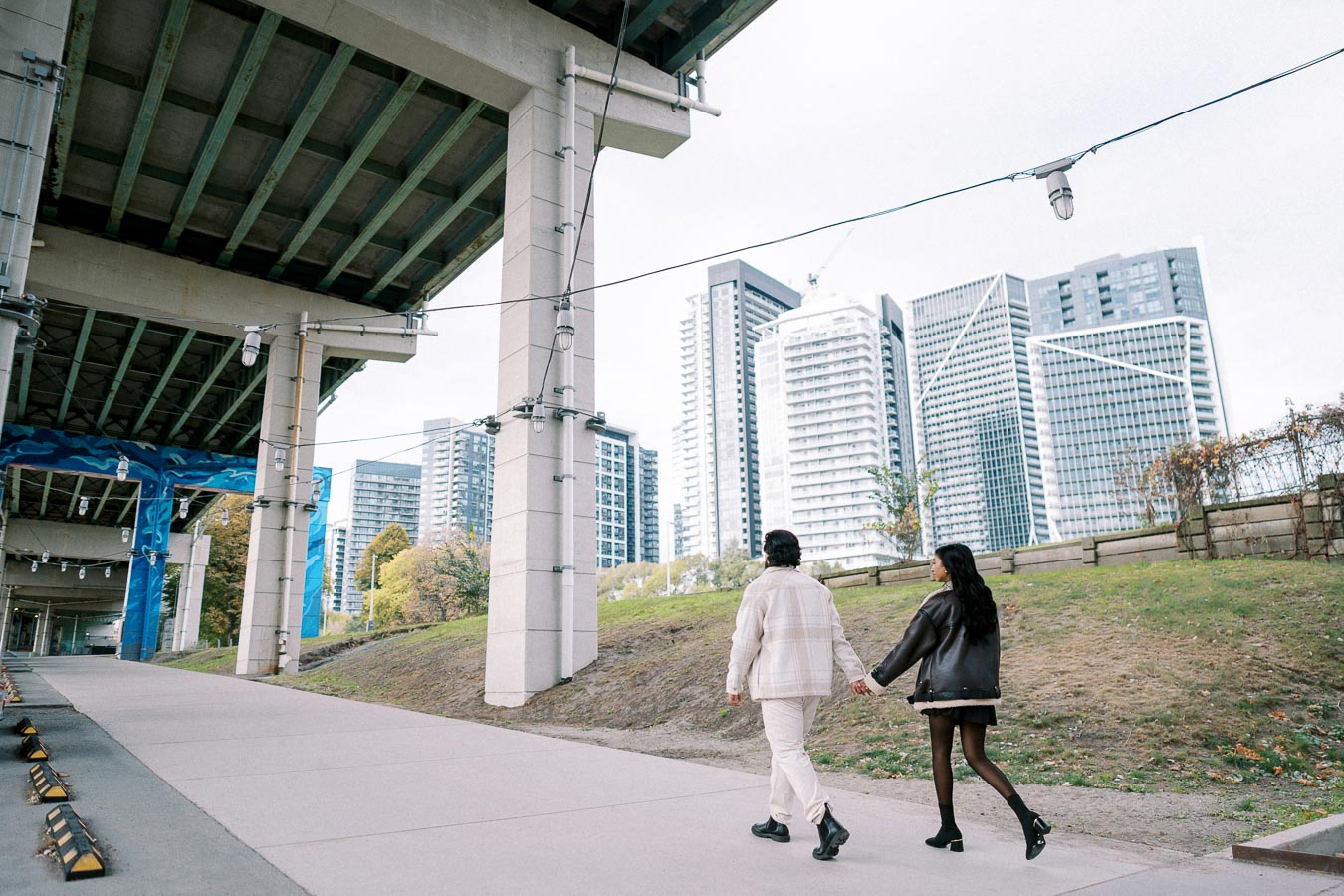 A couple walking hand in hand under an urban bridge with modern skyscrapers in the background.