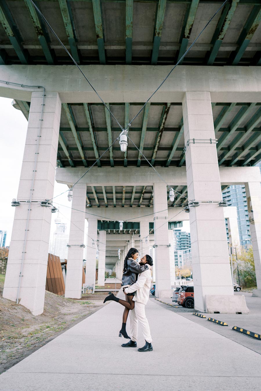 A couple embracing joyfully under an urban bridge, surrounded by concrete columns and city buildings, symbolizing love and connection in an urban environment.