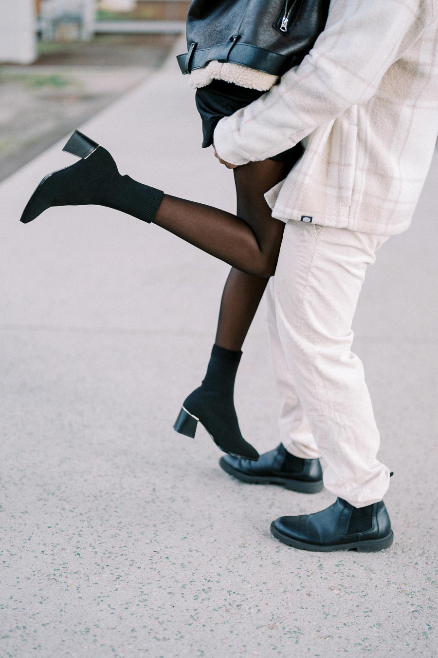A close-up of a stylish couple playfully embracing on a sidewalk, with one person wearing black heeled ankle boots, sheer tights, and a leather jacket, while the other is in light-colored pants with black dress shoes, creating a fashionable and romantic urban scene.
