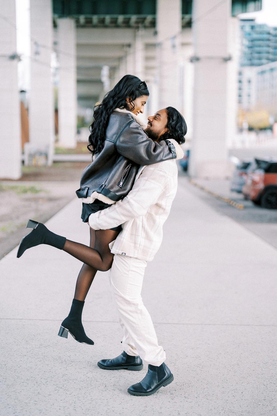 A joyous couple embrace, with the woman in a stylish leather jacket and black heeled boots being lifted by the man in a plaid jacket and light trousers, under a city bridge.