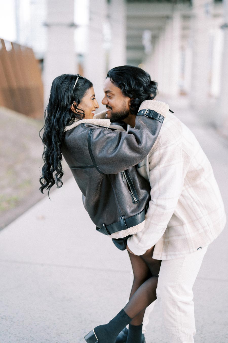 A couple wearing cozy jackets sharing a joyful moment outdoors, engaging in a playful embrace on a city pathway.