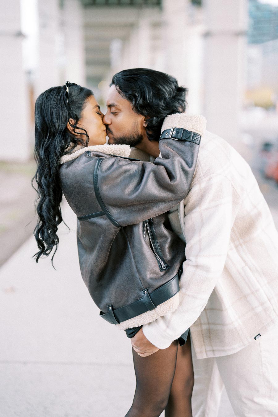 A couple wearing winter coats kissing passionately under an urban bridge, capturing a romantic moment in an outdoor setting.