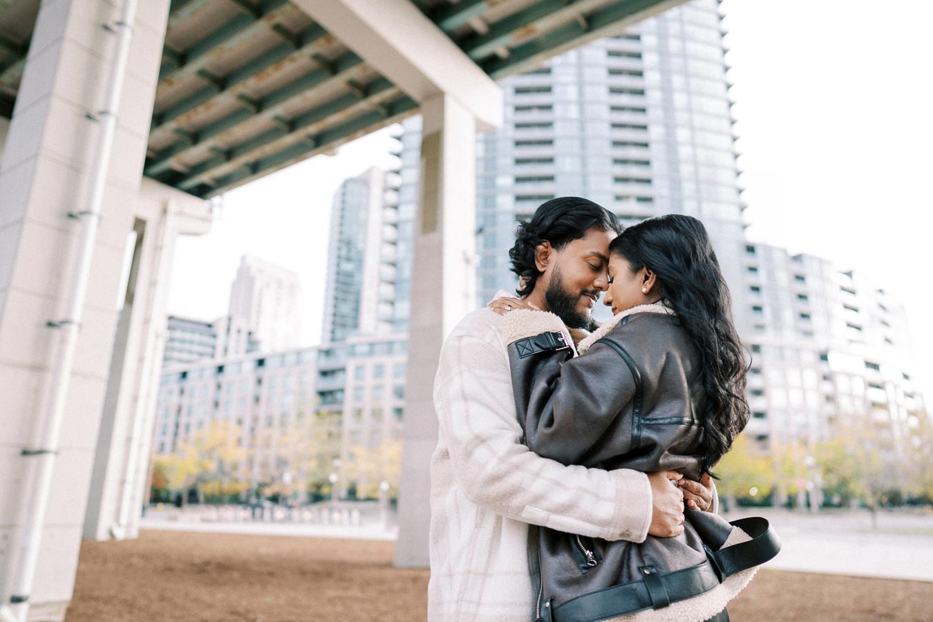 Couple embracing under a modern urban bridge with tall city buildings in the background.