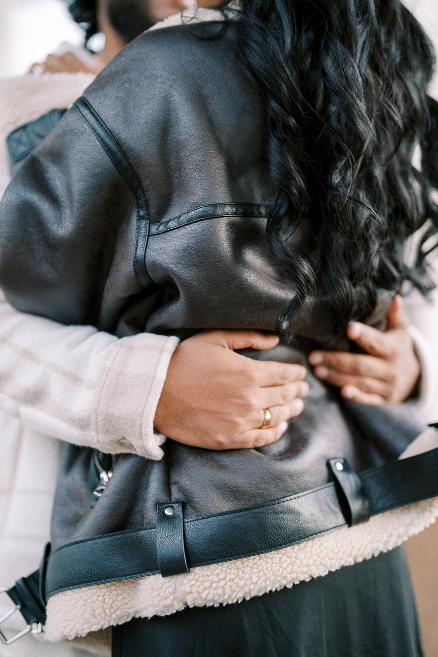 A couple embracing, focusing on their intertwined arms and hands. Both are wearing cozy winter jackets, with one featuring faux fur lining. The woman's long, dark hair cascades down her back. The man wears a ring, suggesting a possible engagement or marriage. The scene conveys warmth and affection.