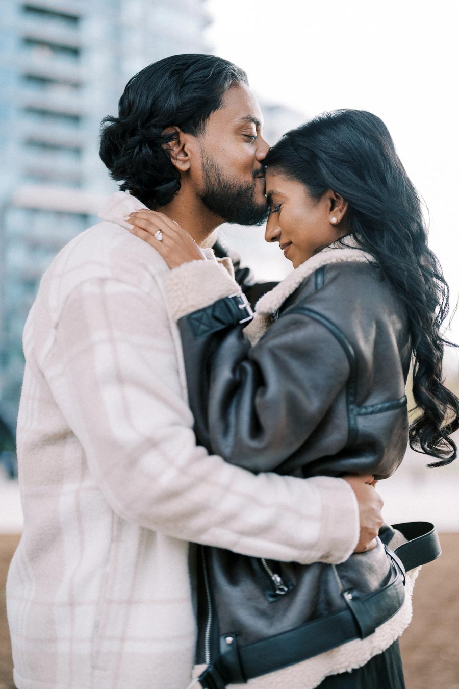 A couple embracing warmly in stylish outerwear, standing closely together with affectionate expressions, against an urban background.