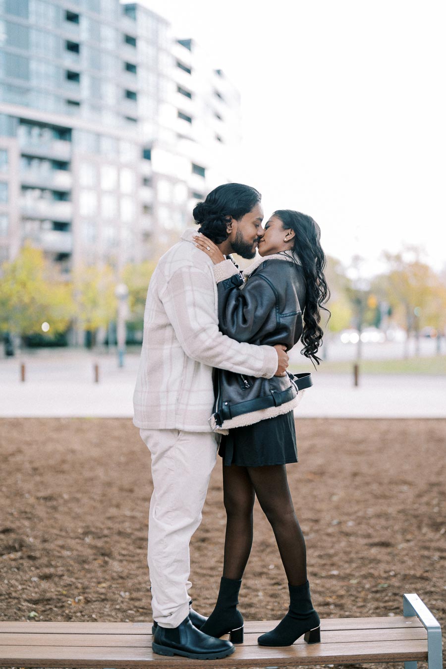 A couple sharing a romantic kiss in a city park, surrounded by modern buildings and autumn trees.