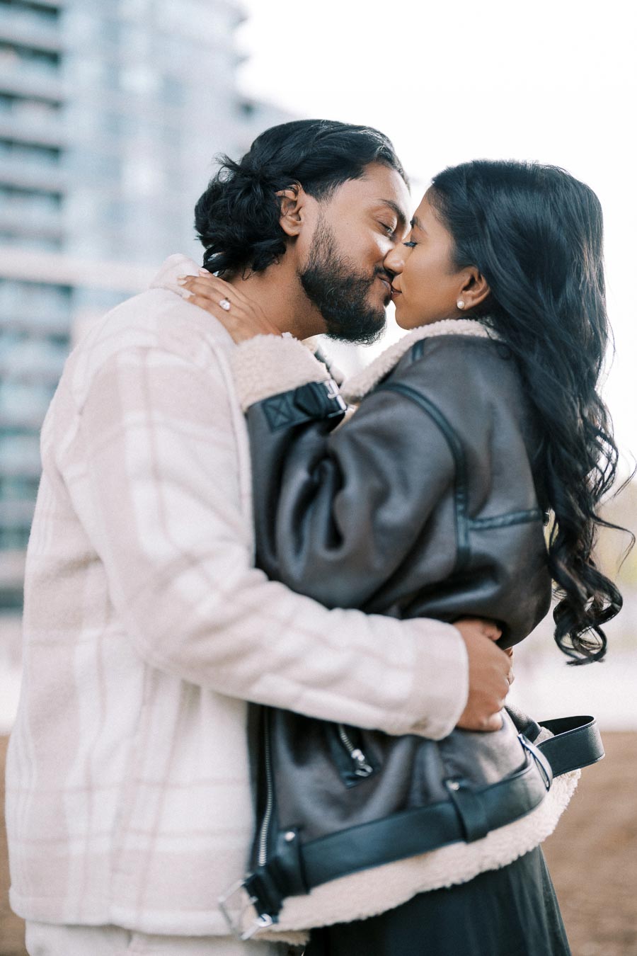 Romantic couple kissing outdoors in cozy winter coats, displaying love and affection amidst an urban backdrop.