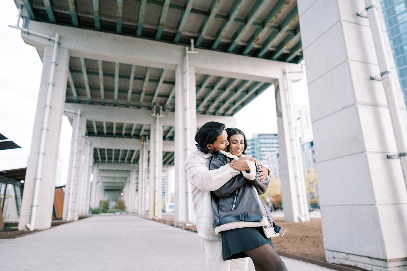 A couple embracing under a modern urban bridge with tall white pillars, capturing a moment of affection in a city setting.