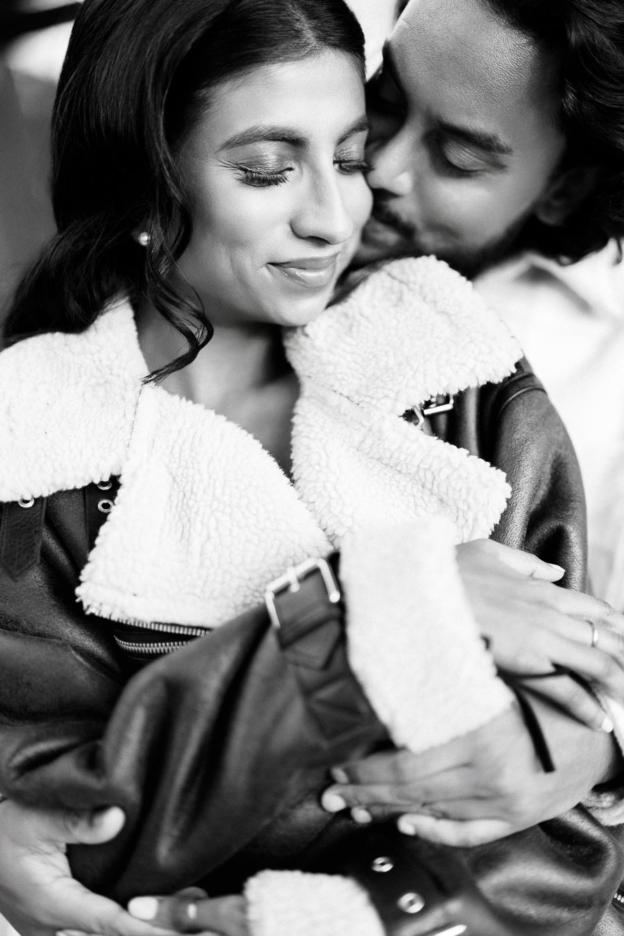 A black and white image of a couple embracing, the woman smiles as she is lovingly held by a man behind her, wearing a cozy shearling-lined jacket.