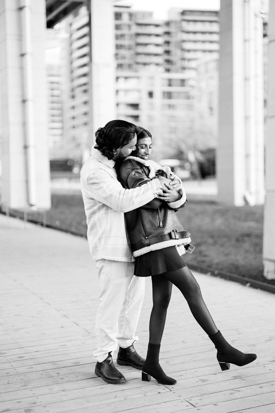 A black and white image of a couple embracing affectionately on a wooden pathway, with urban architecture in the background. The woman is wearing a leather jacket and the man is dressed in light-colored clothing.