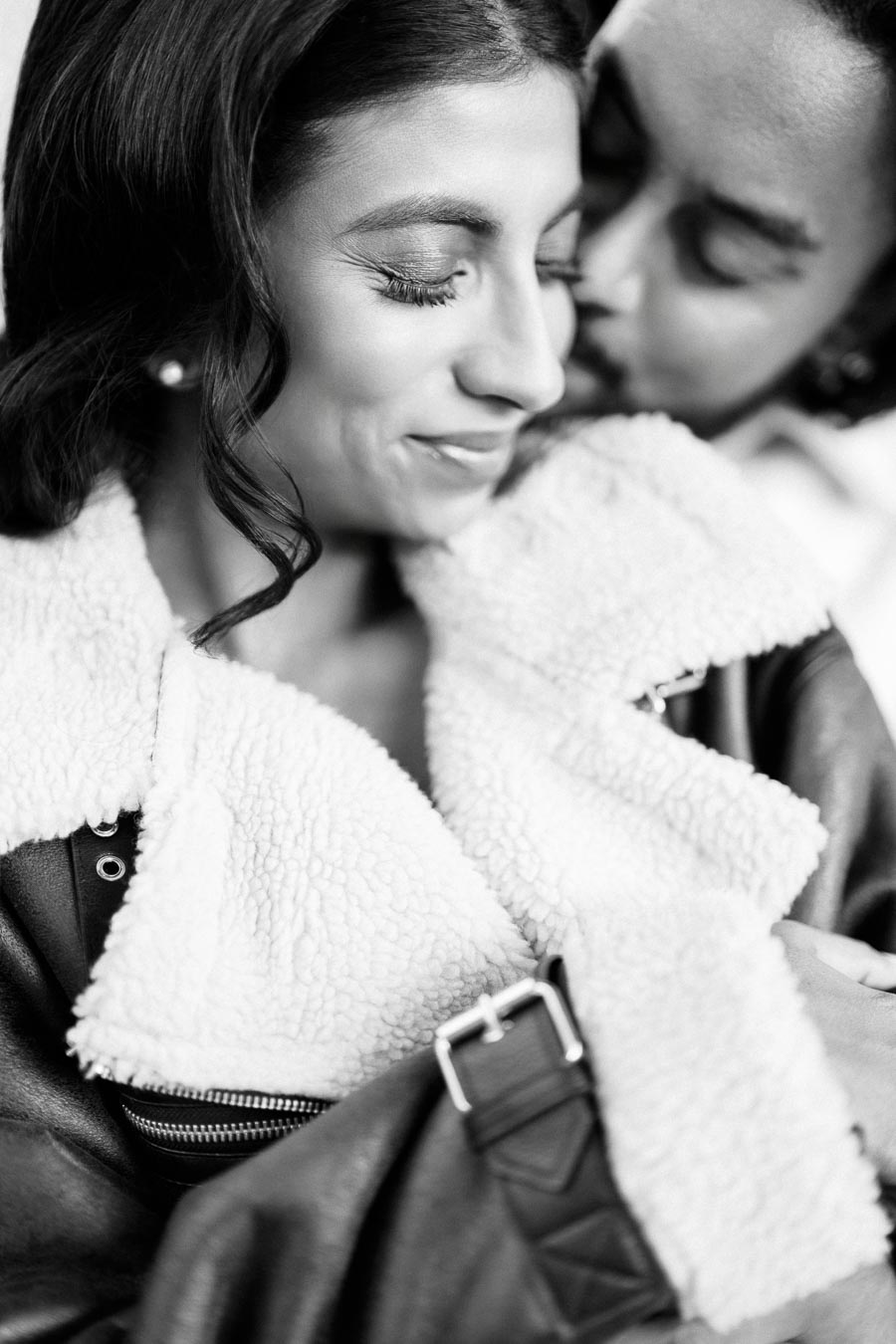 Black and white photo of a couple embracing, with focus on woman smiling and man gently kissing her cheek, both in winter attire with shearling details.