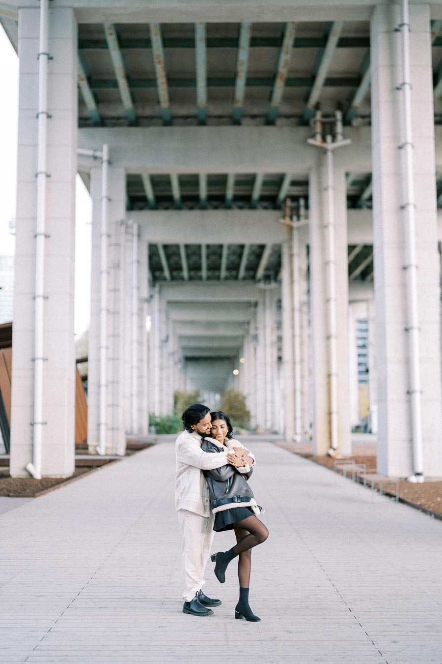 A couple embraces lovingly under a modern urban bridge, with towering concrete pillars stretching into the distance. The serene moment captures connection and affection in an architectural setting, ideal for a romantic photography concept.