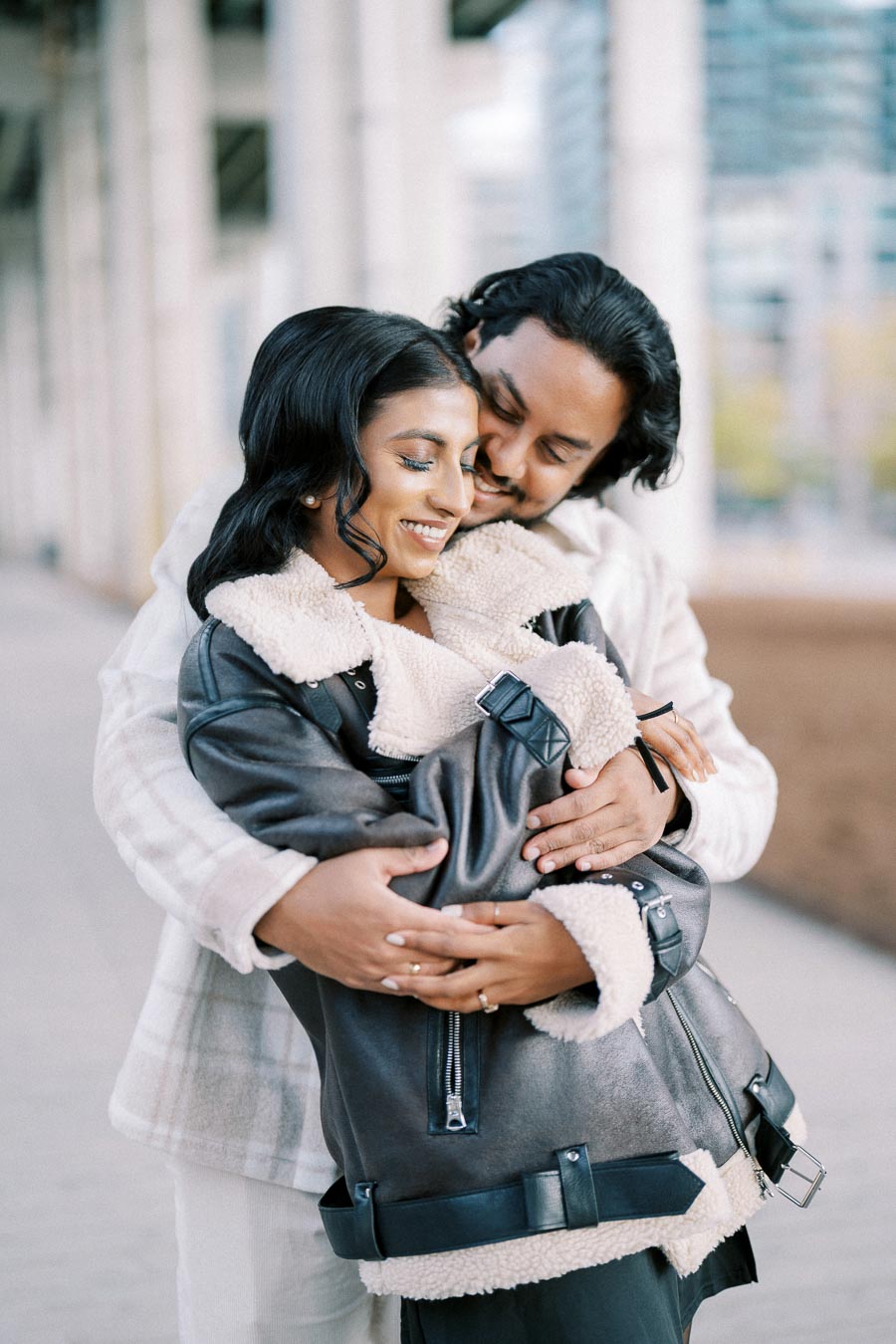 A couple smiling and embracing in a cozy outdoor setting, wearing stylish outfits with layered textures and neutral colors.