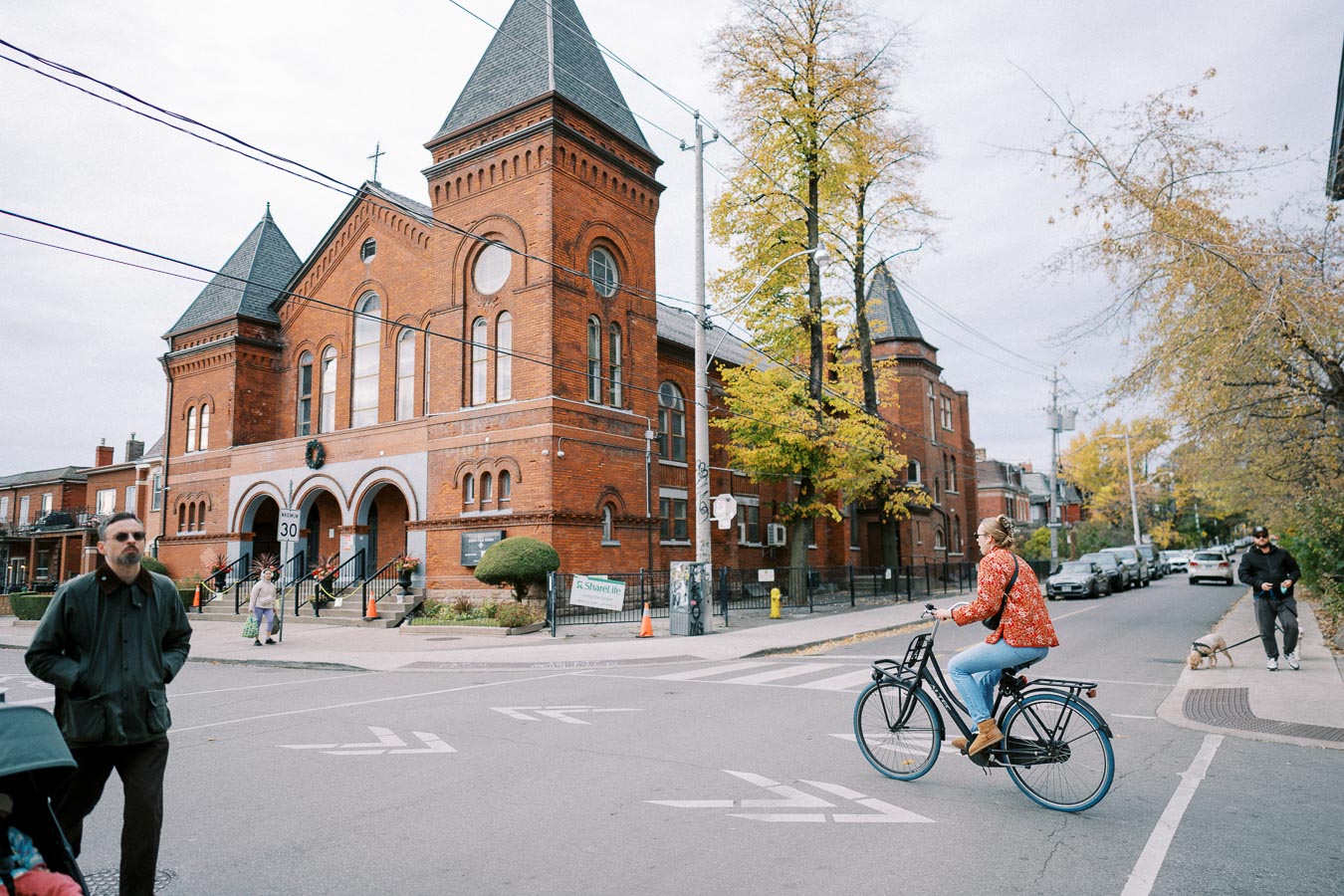 A person bicycling across a street in front of a historic brick church with pointed towers on a cloudy day. People are walking nearby, and autumn trees add color to the urban scene.