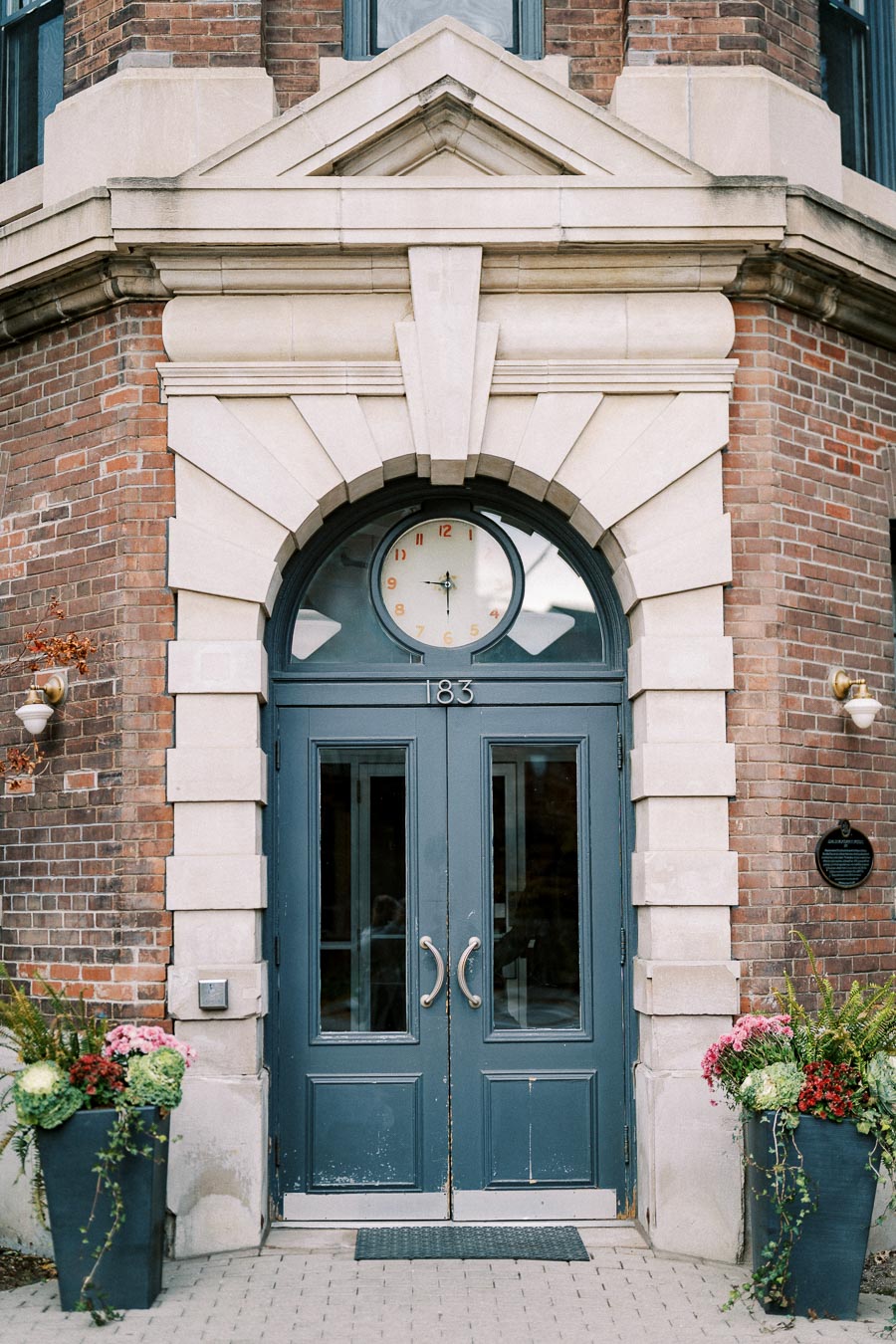 Elegant brick building entrance with blue double doors, ornate stone trim, and a round clock above. Flanked by lush floral arrangements and wall-mounted lamps, displaying architecture at a historical address.
