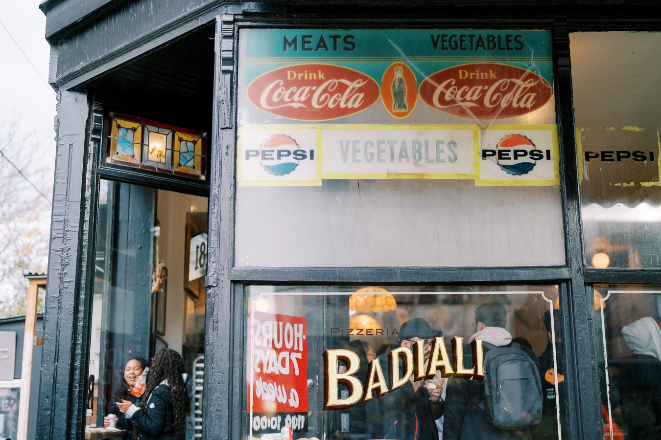 Exterior of a vintage-style pizzeria with Coca-Cola and Pepsi signs, displaying the name 'Badiali' on the window. Two people are seen inside enjoying their time.