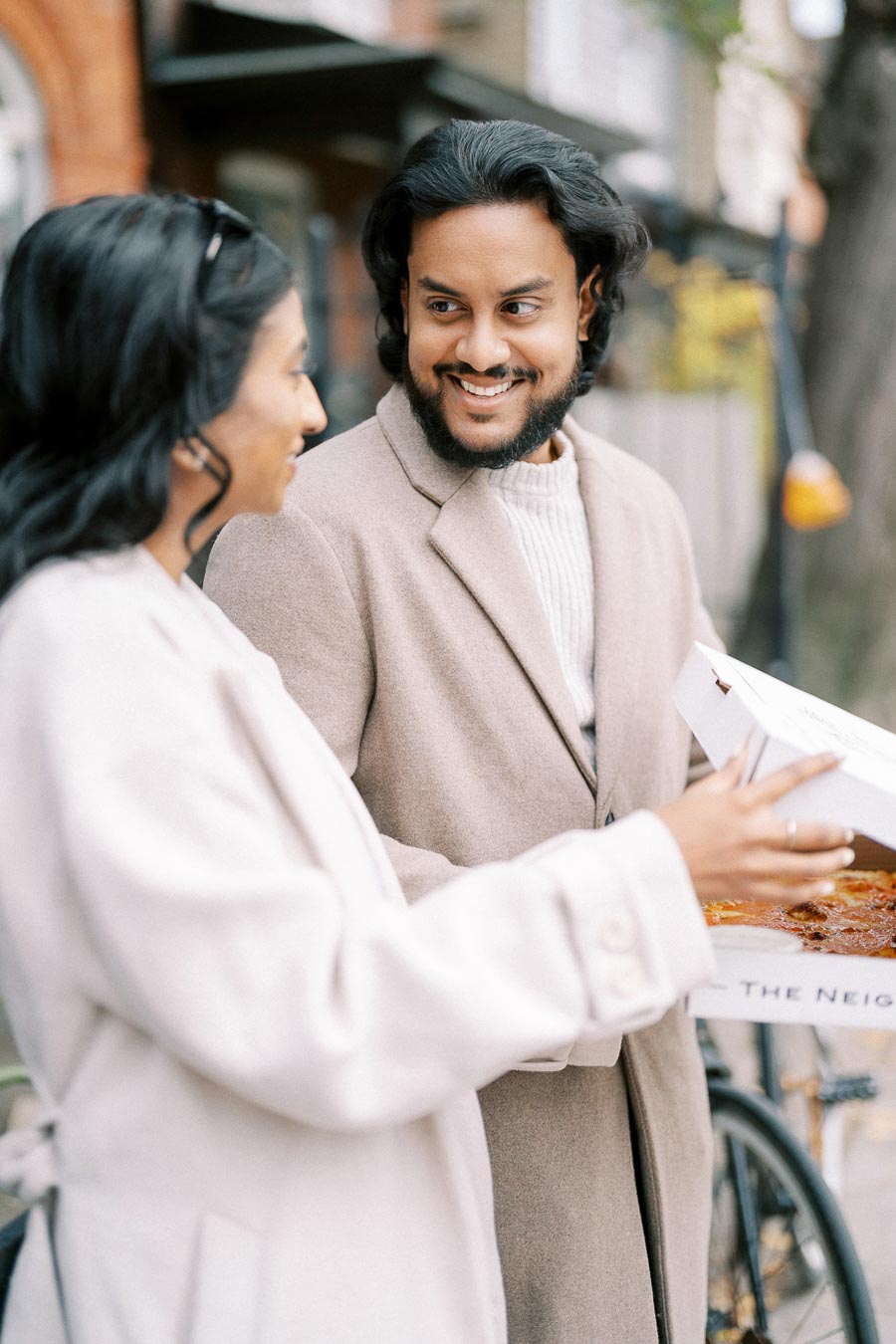 Smiling couple enjoying a pizza outdoors, wearing light beige coats on a casual stroll.