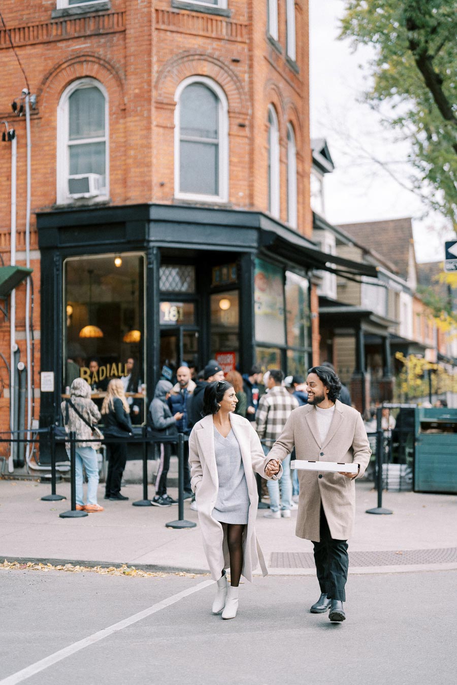 A couple walking hand in hand with a pizza box in a lively urban street, featuring a line of people outside a vintage brick building café.