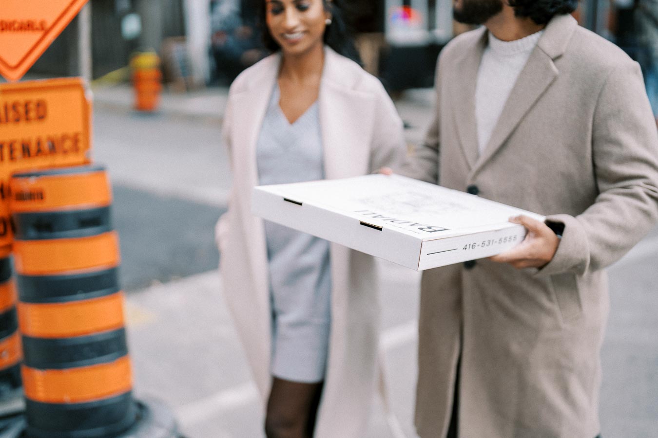 Couple walking on the street with a pizza box, both dressed in beige coats, passing by roadwork cones and a maintenance sign.