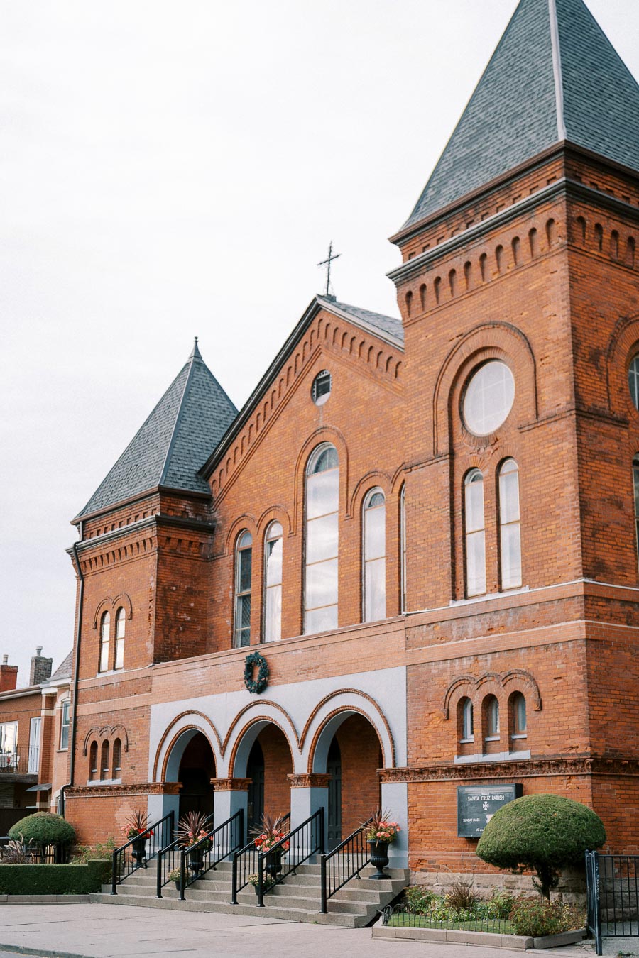 Historic brick church with arched windows and entryways, featuring a cross-topped steeple and landscaped surroundings.