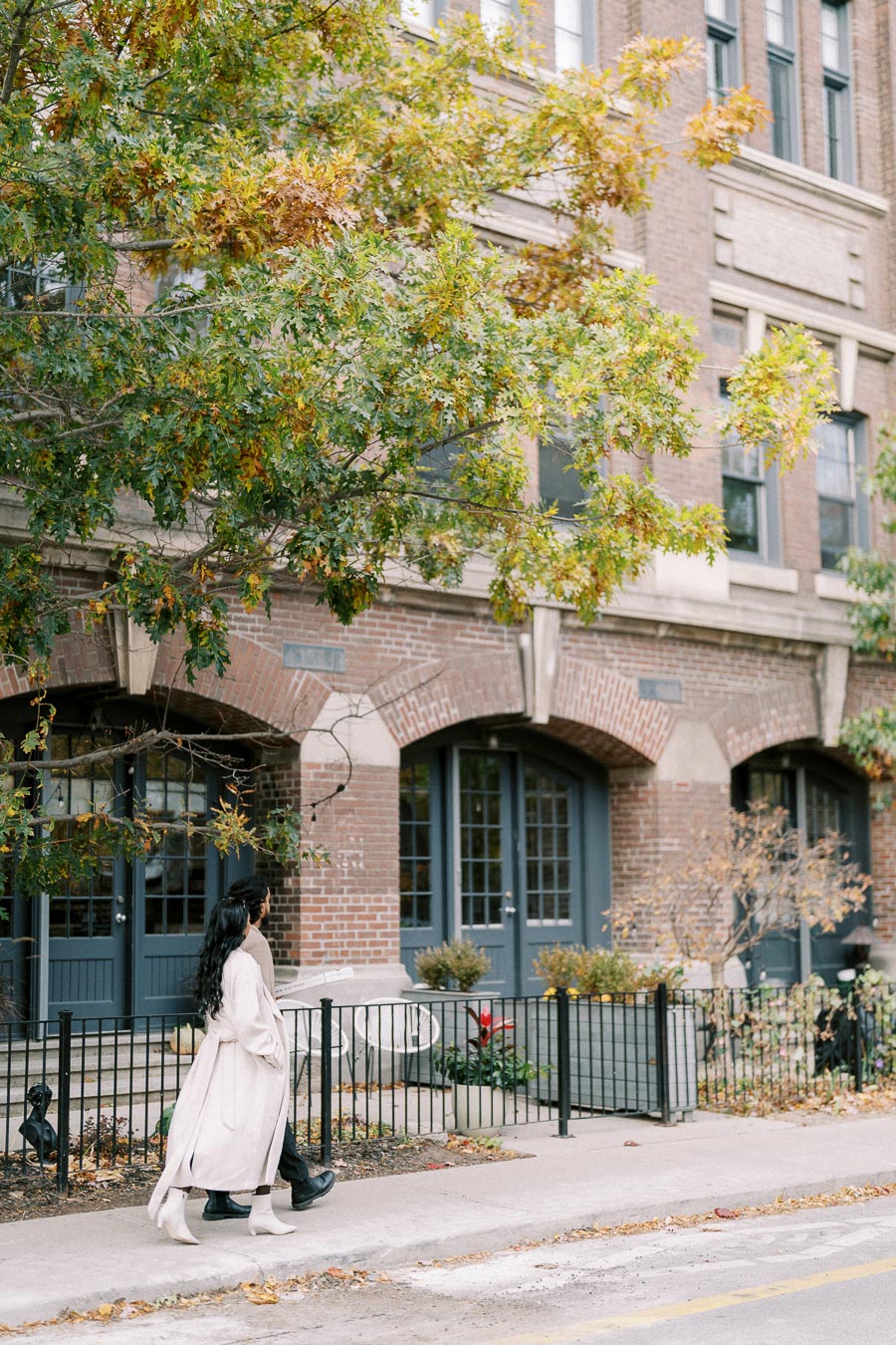 Two people walking on a sidewalk past a brick building with arched windows, surrounded by autumnal trees and light greenery.