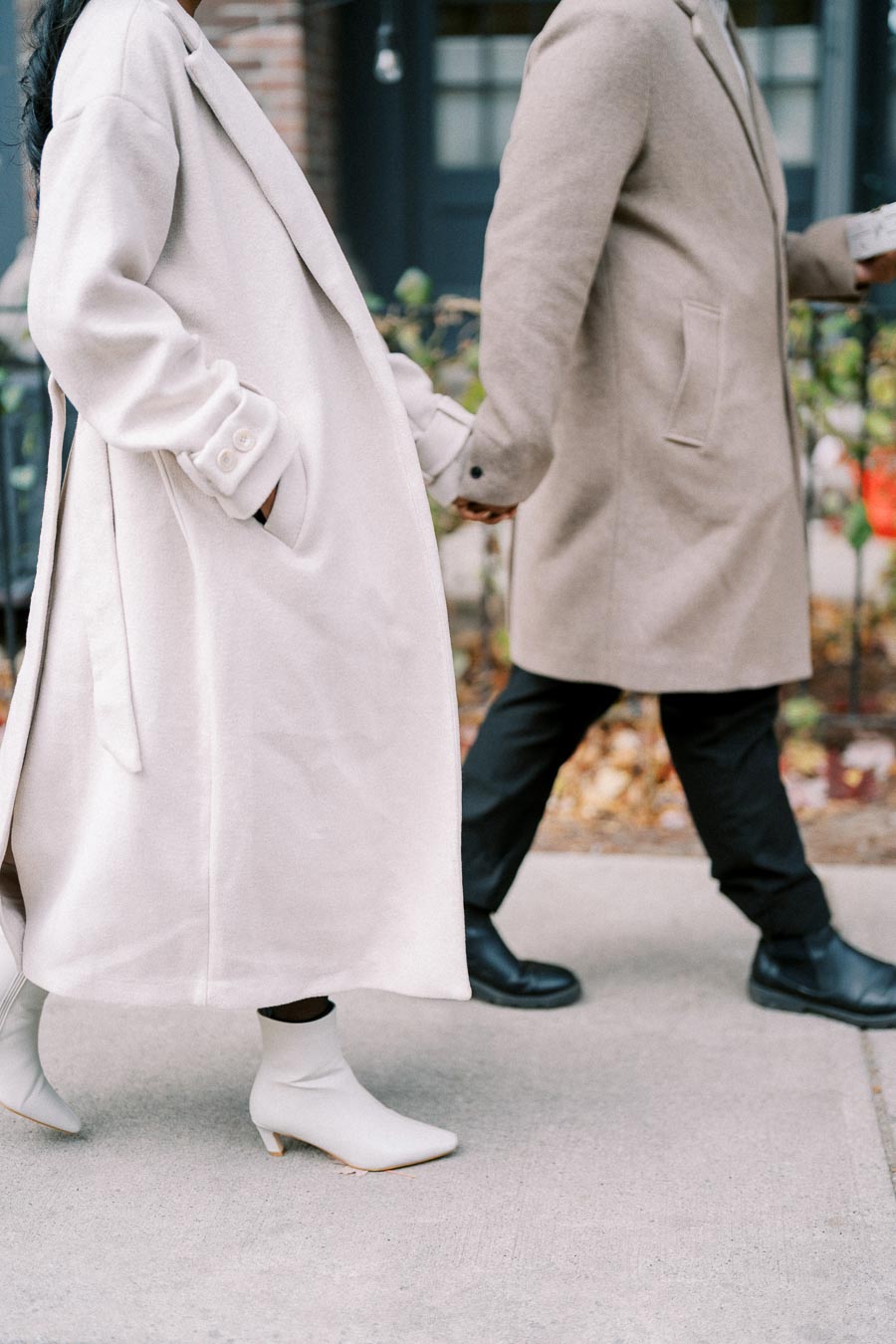 Couple walking hand in hand wearing stylish long beige coats and boots on a city sidewalk.