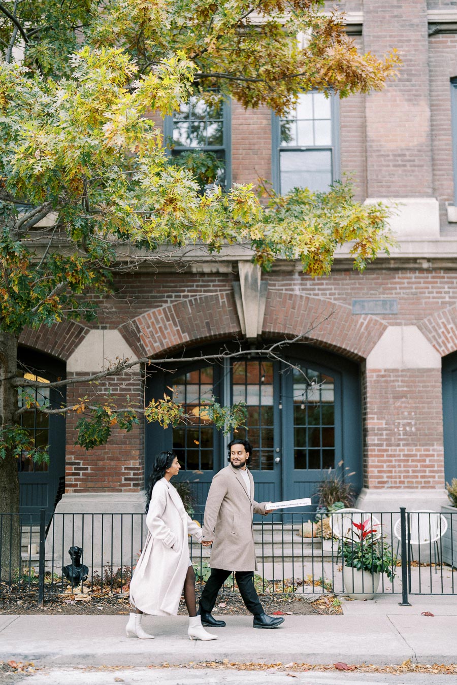 A stylish couple walks hand in hand along a city sidewalk, framed by autumn trees and historic brick architecture, exuding elegance and warmth.